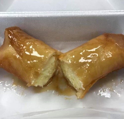 Two golden-brown spring rolls filled with a creamy filling, drizzled with a sweet glaze, on a white paper liner.