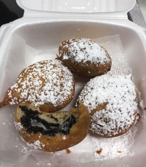 Four powdered sugar-dusted pastries in a takeout container, one cut open to reveal a dark filling.