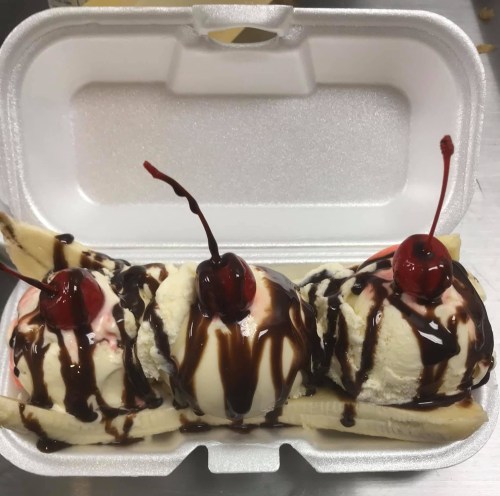 A banana split with three scoops of ice cream, chocolate syrup, and cherries, served in a takeout container.