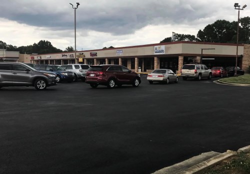 A parking lot with several cars in front of a shopping plaza featuring various storefronts under a cloudy sky.