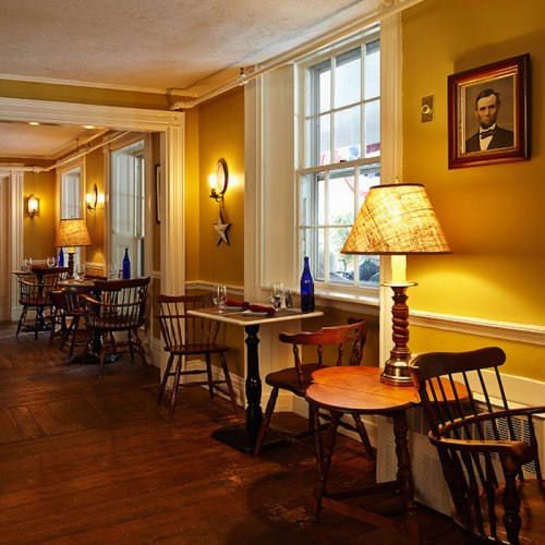 Cozy restaurant interior with wooden tables, chairs, warm lighting, and a portrait on the wall.