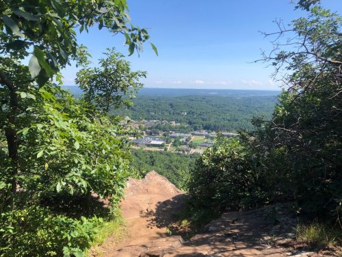 A scenic view from a rocky overlook, surrounded by trees, showcasing a valley and distant hills under a clear blue sky.