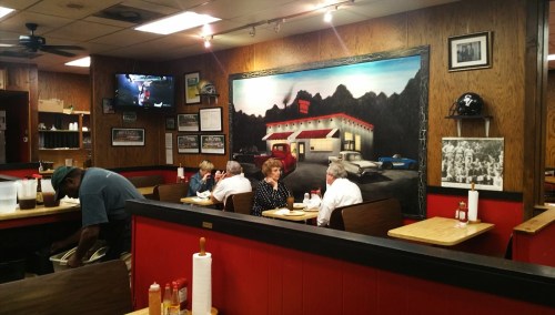 A cozy diner interior with wooden walls, a mural, and patrons enjoying meals at tables. A staff member is in the background.