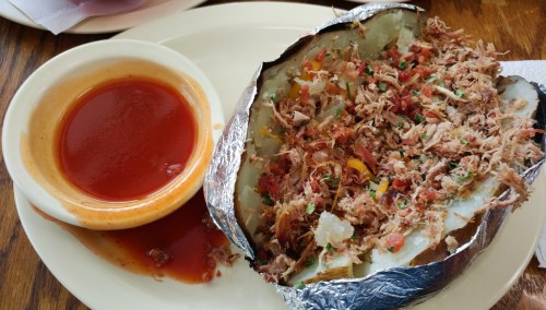 A loaded baked potato topped with shredded meat, tomatoes, and herbs, served with a side of sauce.