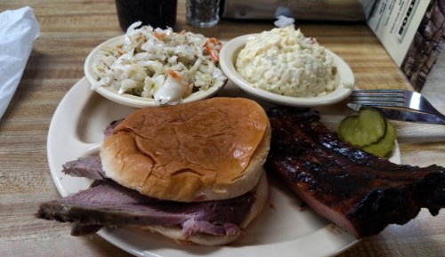 A plate featuring a beef sandwich, ribs, coleslaw, and potato salad, with pickles on the side.