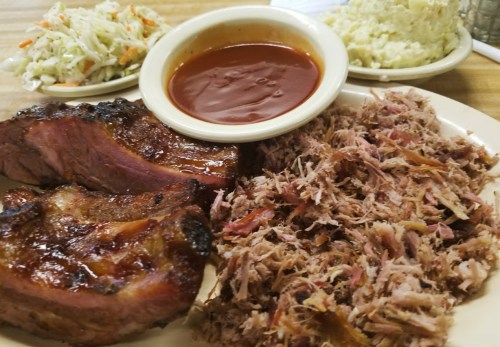 A plate featuring pulled pork, smoked ribs, coleslaw, and a side of barbecue sauce.