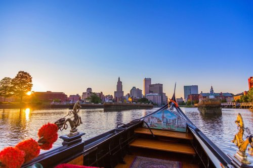 View from a boat on a river at sunset, with a city skyline in the background and warm colors in the sky.