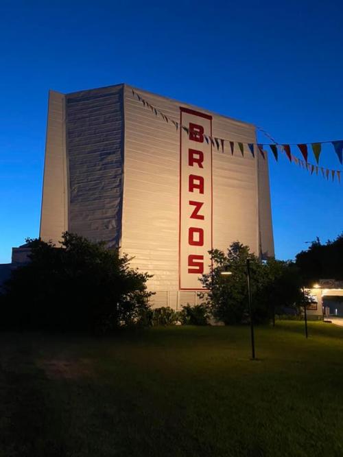 A large building at dusk with "BRAZOS" prominently displayed, surrounded by greenery and colorful flags.