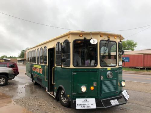 A green and cream vintage-style shuttle bus parked on a street, with a sign reading "Complimentary Shuttle."