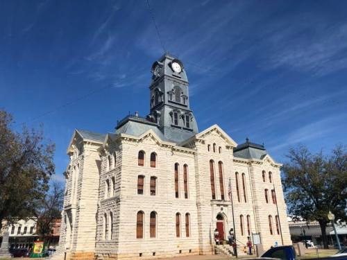 Historic stone courthouse with a clock tower, set against a blue sky with scattered clouds. Trees line the foreground.