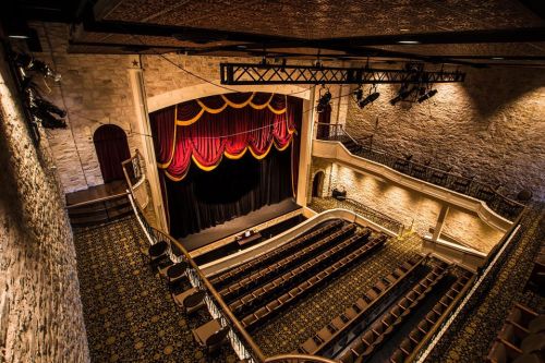 A grand theater interior with a stage, red curtains, and rows of empty seats, featuring stone walls and ornate details.