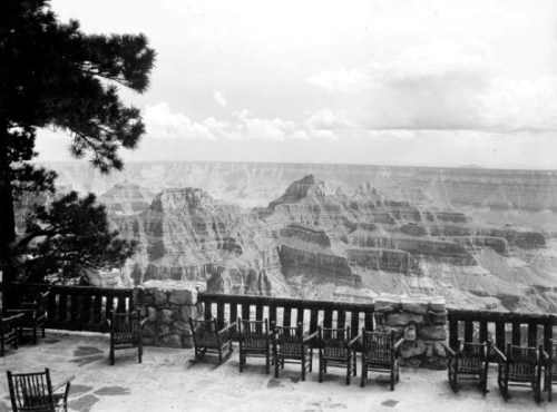Black and white image of a scenic view of the Grand Canyon from a stone terrace with wooden chairs.