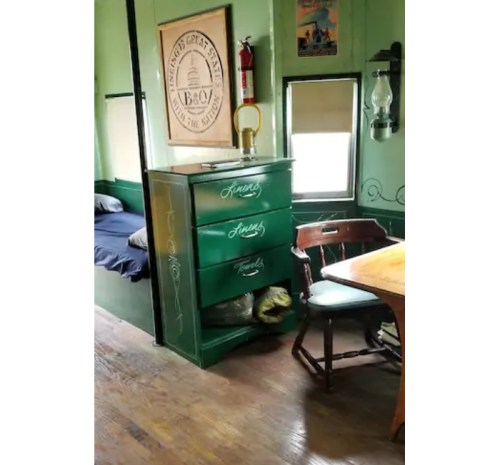 A cozy room featuring a green dresser, wooden table, and a bed, with vintage decor and warm lighting.