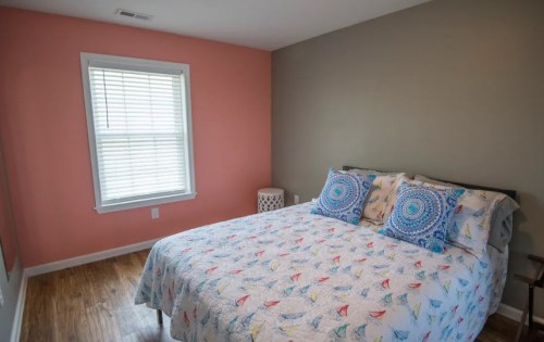 Cozy bedroom with a double bed, colorful bedding, and a window with blinds, featuring a peach and gray wall.
