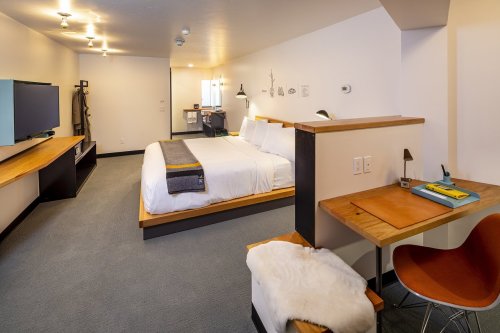 Modern hotel room featuring a large bed, desk, TV, and minimalist decor with wooden accents.