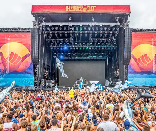 A lively crowd at a music festival, with a large stage and colorful banners, enjoying the performance.