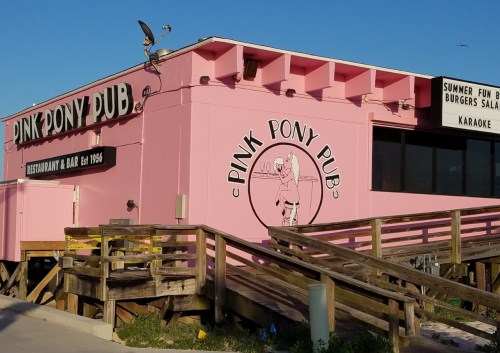 A pink building with "Pink Pony Pub" signage, featuring a restaurant and bar, with a wooden ramp leading up to it.