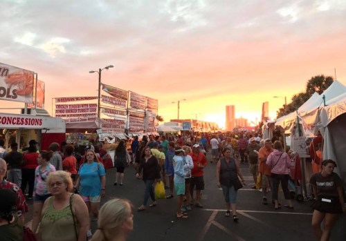 Crowd at a fair with food stalls under a vibrant sunset sky, creating a lively and festive atmosphere.