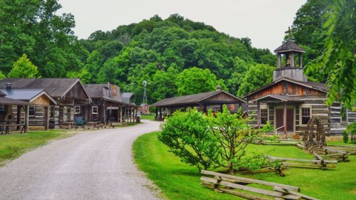 A scenic view of a rustic village with wooden buildings, a gravel path, and lush green trees in the background.