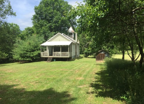 A white house with a porch and bell tower, surrounded by trees and a grassy yard, with a small shed nearby.