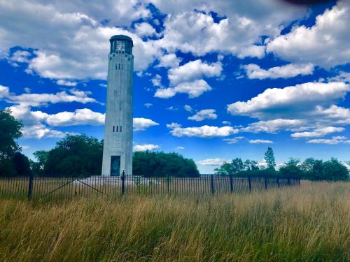 A tall, white tower stands amidst tall grass and trees under a blue sky with fluffy clouds. A fence surrounds the area.