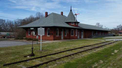 Historic train station with a red brick exterior, green roof, and nearby railroad tracks under a clear blue sky.