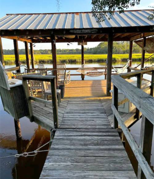 A wooden dock leads to a gazebo overlooking a calm marsh, with chairs and a hammock for relaxation.