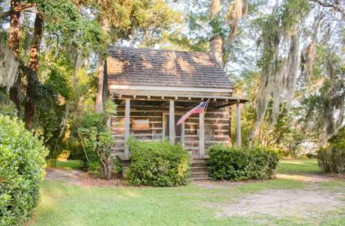 A rustic log cabin surrounded by greenery and trees, featuring a porch with rocking chairs and an American flag.
