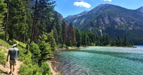 A scenic view of a lake surrounded by mountains and trees, with two hikers walking along a path by the water.