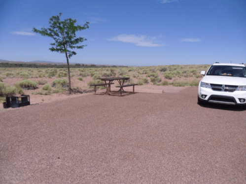 A picnic table and a white car are parked in a sandy area with sparse vegetation and a clear blue sky.