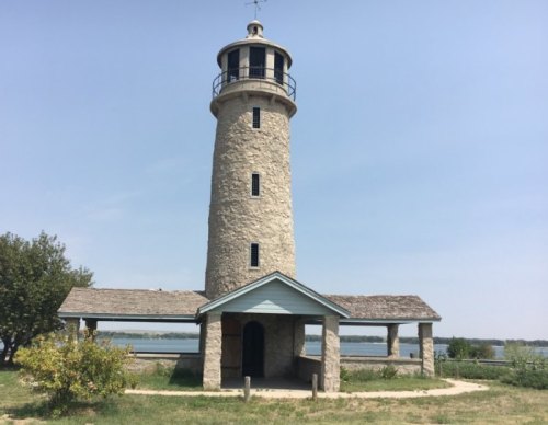 A tall stone lighthouse with a black railing, surrounded by greenery and overlooking a calm body of water.