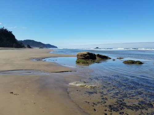 A serene beach scene with gentle waves, rocky formations, and a clear blue sky in the background.