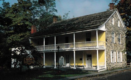 Historic two-story stone house with yellow siding and a porch, surrounded by trees and a historical marker.