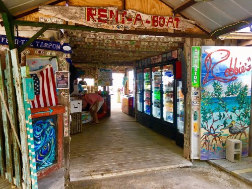 A rustic boat rental shop with colorful murals, dollar bills on the walls, and a cooler filled with drinks.