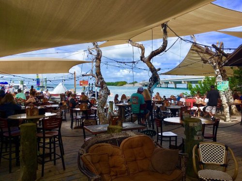 A lively outdoor restaurant by the water, featuring shaded seating and patrons enjoying the view.