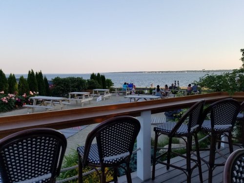 View of a waterfront patio with empty chairs, picnic tables, and people enjoying the scenery at sunset.