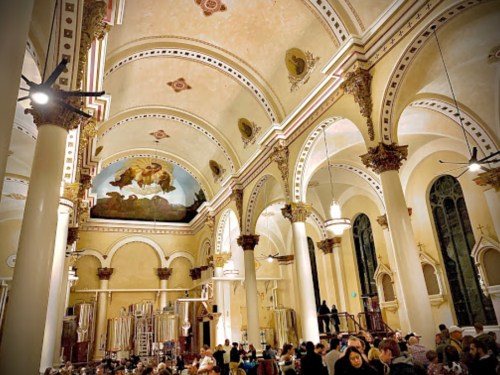 Interior of a grand church with high arches, ornate columns, and a large mural, filled with people dining below.