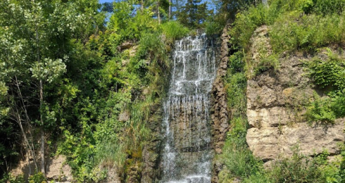 A cascading waterfall surrounded by lush greenery and rocky cliffs under a clear blue sky.