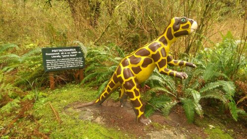 A colorful dinosaur statue, Psittacosaurus, stands among ferns with an informational sign nearby in a forest setting.