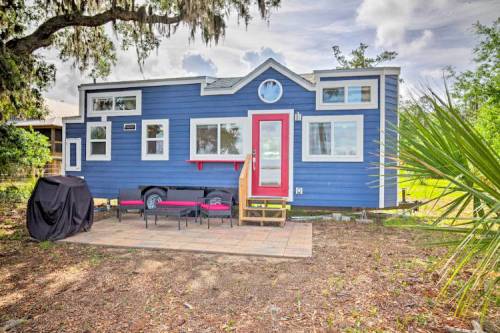 A blue tiny house with large windows, a red door, and a patio area surrounded by greenery and trees.