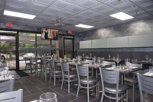 Modern restaurant interior with gray decor, tables set for dining, and a TV displaying a scene on the wall.