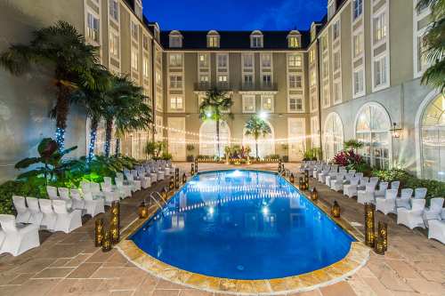 A beautifully lit pool area surrounded by palm trees and chairs, with string lights creating a festive atmosphere at dusk.