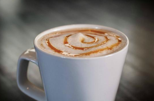 A close-up of a white mug filled with coffee topped with frothy milk and a caramel swirl.