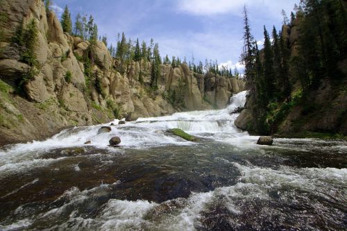 A scenic waterfall cascades over rocky cliffs, surrounded by lush greenery and tall trees under a blue sky.