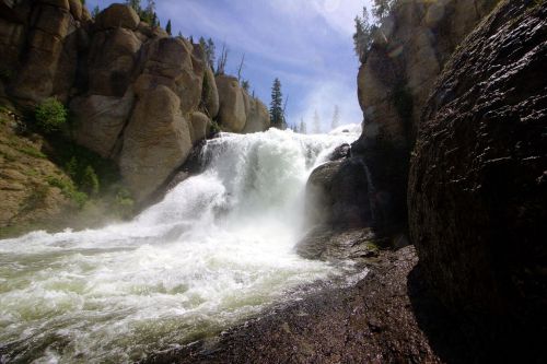 A powerful waterfall cascades over rocky cliffs, surrounded by lush greenery and tall trees under a bright blue sky.
