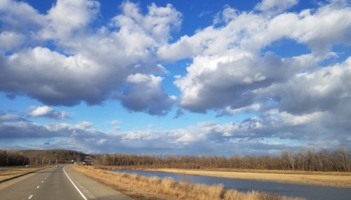 A scenic view of a highway alongside a river, with a blue sky filled with fluffy clouds and bare trees in the background.