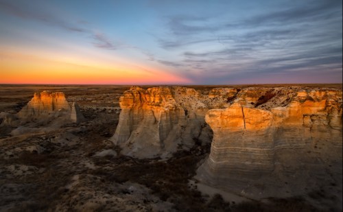 Sunset over rugged rock formations, casting warm light on cliffs against a colorful sky in a vast landscape.