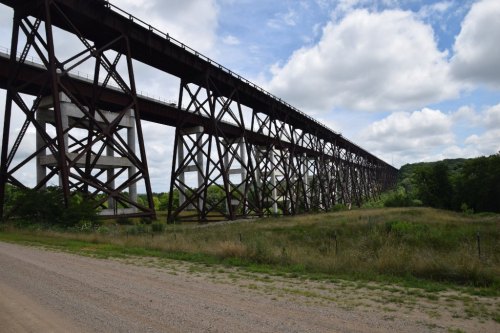 A tall, rust-colored railway bridge spans a green landscape under a partly cloudy sky.