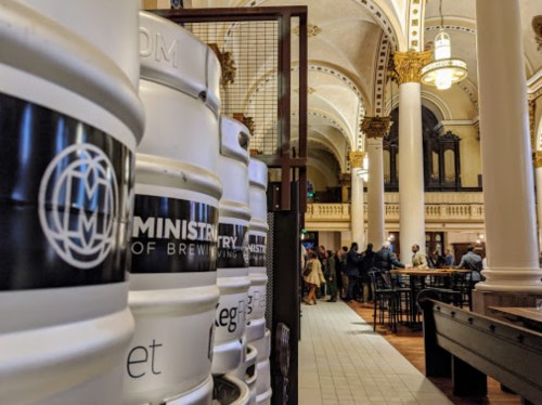 Interior of a brewery with barrels in the foreground and people socializing in a grand, historic setting.