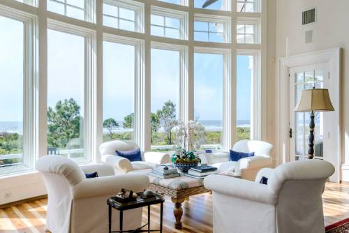 Bright living room with large windows overlooking the ocean, featuring white furniture and a wooden coffee table.